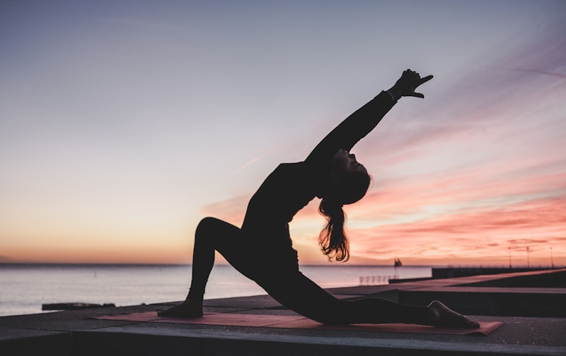 Silhouette of a person in a yoga lunge pose on a mat at sunrise by the water